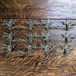May include: A set of eight ornate brass drawer pulls with a floral design. The pulls are arranged in three rows on a wooden surface.