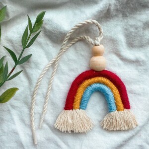 May include: A colorful rainbow made of yarn with a wooden bead hanging from it. The rainbow is red, orange, yellow, blue, and green. The wooden bead is brown. The rainbow is hanging from a white string.