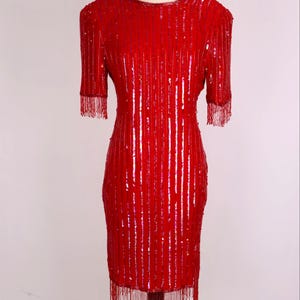 May include: A vibrant red sequined dress with short sleeves and fringe detailing. The dress is knee-length and features vertical sequin stripes. The sleeves and hemline are adorned with matching red fringe. The dress is displayed on a mannequin.