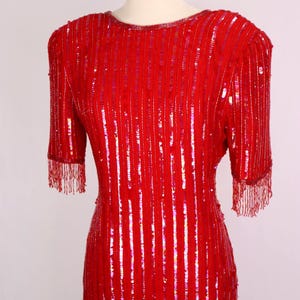 May include: A vibrant red sequined dress with a round neckline and short sleeves. The dress features vertical rows of sequins and beaded fringe at the sleeve ends. The dress is displayed on a mannequin.