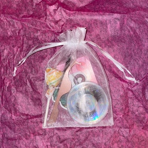 May include: A white organza bag filled with lash supplies, including a pair of false eyelashes, a lash applicator, and a lollipop.