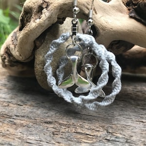 May include: A pair of silver whale tail earrings with a gray crocheted rope design. The earrings are hanging from a wooden surface.