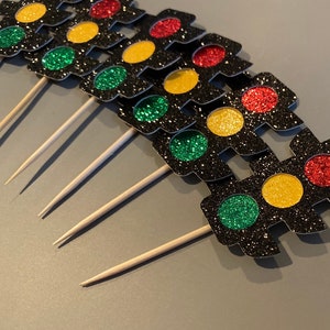Op de afbeelding: Een set van zes zwarte glitter verkeerslicht cupcake toppers met rode, gele en groene glitter cirkels. De toppers zijn bevestigd aan houten spiesjes.