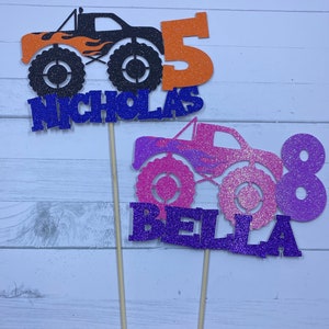 May include: Two monster truck cake toppers, one black with orange flames and the number 5, and the other pink with purple flames and the number 8. The toppers are decorated with glitter and have the names "Nicholas" and "Bella" written below the trucks.