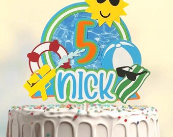 May include: A cake topper with a summer theme, featuring a yellow sun wearing sunglasses, a blue and orange number 5, and the name "Nick". Other elements include a beach ball, a life preserver, and a water gun.