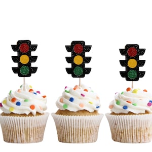 Op de afbeelding: Drie cupcakes met witte frosting en kleurrijke sprinkles, bedekt met zwarte, rode, gele en groene verkeerslicht cupcake toppers.