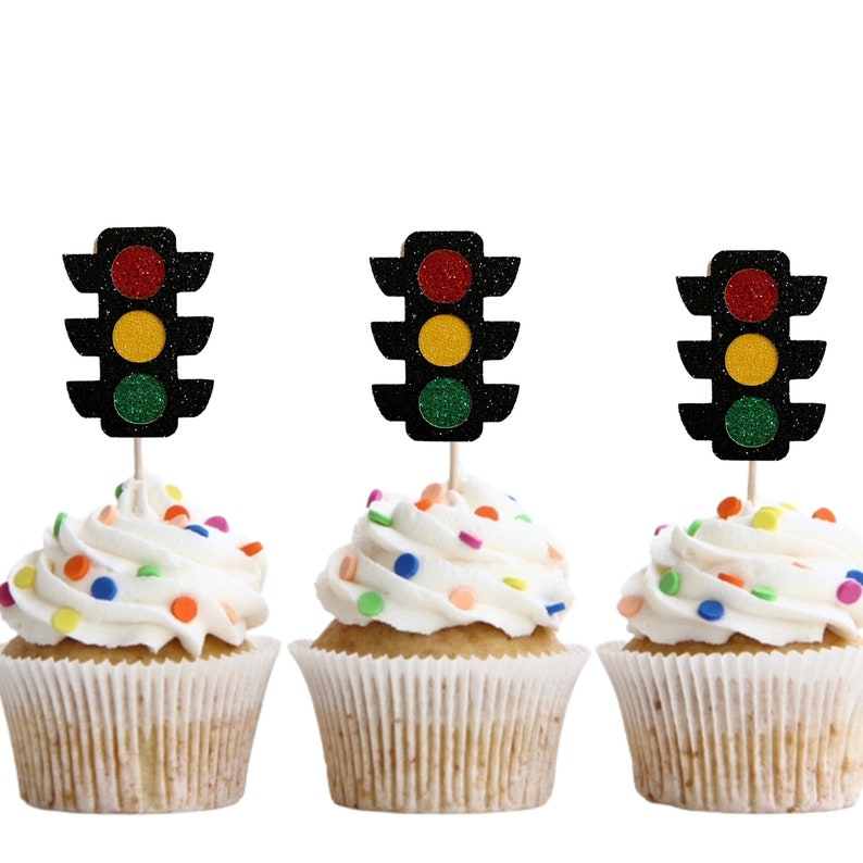 Op de afbeelding: Drie cupcakes met witte frosting en kleurrijke sprinkles, bedekt met zwarte, rode, gele en groene verkeerslicht cupcake toppers.
