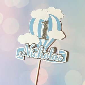May include: Blue and white hot air balloon cake topper with the number 1 and the name Nicholas in silver glitter.