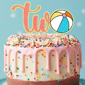 May include: A two-tiered birthday cake with pink frosting drips and colorful sprinkles. A glittery pink "two" cake topper with a yellow outline and a beach ball illustration adds a festive touch. The cake sits on a light blue background.