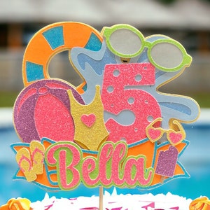 May include: A colorful cake topper with a beach theme. It features a pink and purple beach ball, a blue and green life preserver, a gold and pink swimsuit, a blue and pink number 5, and the name "Bella" in pink and green letters.