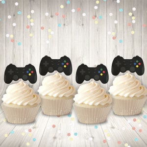 May include: Four cupcakes decorated with black and white video game controller toppers. The cupcakes are white with white cupcake liners. The background is a light woodgrain with colorful confetti.