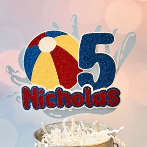 May include: A blue and red beach ball with a yellow center, a blue glitter number 5, and the name Nicholas in red glitter letters. The design is on a white background with a blue glitter water splash design.