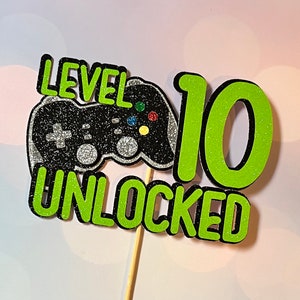K&ouml;nnte beinhalten: Gr&uuml;ner und schwarzer Tortendekor mit dem Text "Level 10 Unlocked" und einem schwarzen und silbernen Videospiel-Controller.