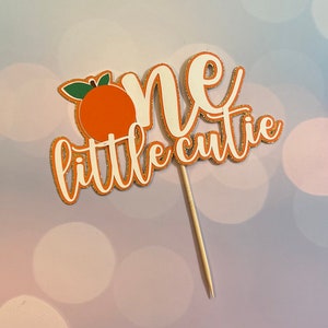 May include: Orange and white cake topper with gold glitter accents. The topper says "one little cutie" and features a small orange with green leaves.