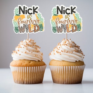 May include: Two cupcakes with white frosting and gold sprinkles topped with a jungle animal themed cupcake topper that says "Nick is forever wild".