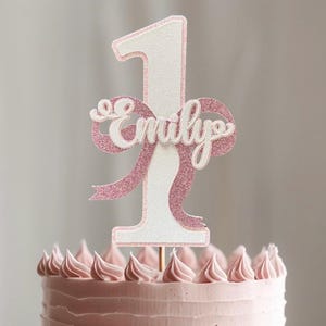 May include: A pink and white cake topper in the shape of the number 1, with a pink glitter bow and the name "Emily" in white script. The topper is placed on a pink frosted cake.