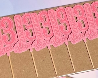 May include: Pink cupcake toppers with the text "Little Miss Three" on each topper. The toppers are on wooden sticks.
