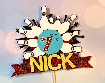 May include: A red and gold glitter cake topper with a bowling ball and pins design. The bowling ball is blue with white stars and the number 7. The topper also includes the name "NICK" in gold glitter.