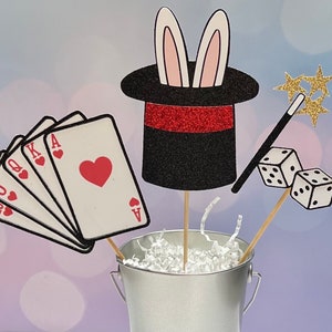 May include: A magic themed centerpiece with a black top hat with bunny ears, playing cards, dice, and a magic wand. The items are arranged in a silver bucket with white shredded paper.