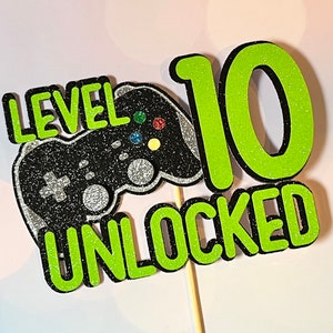 May include: Green and black cake topper with a video game controller and the text "Level 10 Unlocked".