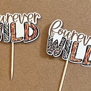 May include: Two white cupcake toppers with the text "forever wild" in brown lettering. The letters are decorated with animal print patterns, including leopard, zebra, giraffe, and tiger.