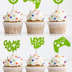 May include: Six cupcakes decorated with green glitter cupcake toppers. The toppers feature a game controller, a power button, the words "Game On", the words "Level Up", and a star.