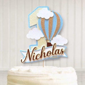 May include: A cake topper featuring a number one with a hot air balloon design in blue, gold, and brown, with white clouds. The name "Nicholas" is written on a banner below the number one. The topper is placed on top of a white frosted cake.