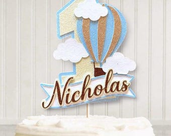Könnte beinhalten: Ein Tortenaufsatz für einen Geburtstag. Er zeigt die Zahl eins, einen blau-gold-braunen Heißluftballon und weiße Wolken. Der Name "Nicholas" steht auf einem Banner. Eine festliche Dekoration für den ersten Geburtstag.