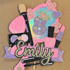 May include: A pink and purple glitter cake topper with a mirror, makeup brushes, and nail polish. The topper has a banner with the name "Emily" in gold glitter.