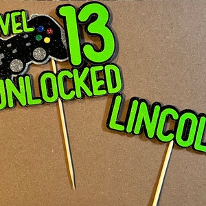 K&ouml;nnte beinhalten: Zwei gr&uuml;ne und schwarze Glitzer-Kuchentopper mit dem Text "Level 13 Unlocked" und "Lincoln".