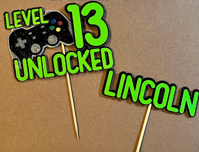 K&ouml;nnte beinhalten: Zwei gr&uuml;ne und schwarze Glitzer-Kuchentopper mit dem Text "Level 13 Unlocked" und "Lincoln".
