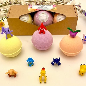 May include: Three spherical bath bombs in yellow, lavender, and peach colours, each topped with a small plastic creature figure. The set is presented in a rectangular cardboard box labelled Children's Bath Bombs With Surprise Toys, accompanied by several loose plastic creature figurines in various colours and shapes.