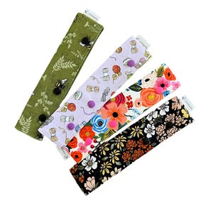 May include: Four fabric face mask extenders with different patterns. The extenders are green with bees, purple with yarn, black with flowers, and black with white flowers. Each extender has two buttons.