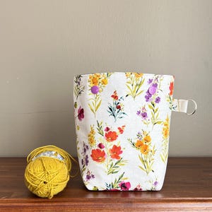 Könnte beinhalten: Eine weiße Garnaufbewahrungstasche mit einem floralen Muster in Rot, Gelb und Lila. Die Tasche hat einen kleinen Griff mit einem Metallring und zwei lila Knopfverschlüssen. Eine Kugel gelbes Garn liegt neben der Tasche.