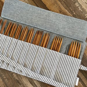 Puede incluir: Un juego de agujas de tejer de madera en un estuche de tela a rayas grises y blancas. Las agujas, de varios tamaños, están dispuestas ordenadamente dentro del estuche abierto. El estuche es de tela gris suave y tiene una solapa.