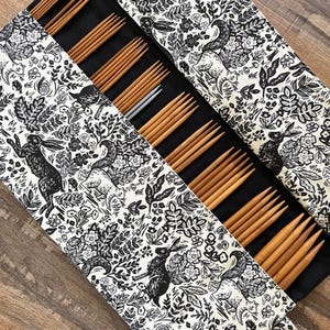 May include: A black and white fabric knitting needle case with a woodland animal print. The case is open, revealing several sets of wooden knitting needles in various sizes. The needles are arranged in rows.