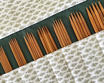 May include: A set of wooden knitting needles organized in a fabric roll-up case. The case features a white fabric with a repeating olive green diamond pattern and a solid dark green interior lining, holding multiple sets of pointed bamboo needles.