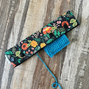 May include: A black floral print yarn needle case with a blue yarn ball. The case features a colorful flower pattern with orange, yellow, and pink blooms. The case is secured with two black buttons. A strand of blue yarn extends from the yarn ball.