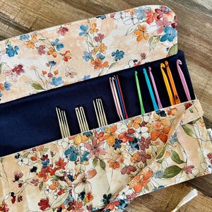 May include: A fabric roll-up organizer featuring a floral pattern in shades of orange, blue, and pink on a cream background. The interior is dark blue and holds several metal knitting needles and colorful crochet hooks in various sizes.