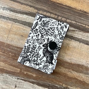 May include: A black and white fabric wallet featuring a woodland design with a rabbit, flowers, and foliage. The wallet has a black button closure. The design is printed on a white background.