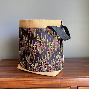 Può includere: Una borsa tote cilindrica con un motivo floreale nero, con fiori rosa, gialli e bianchi. La parte superiore e inferiore sono beige, con un cinturino nero. La borsa è appoggiata su una superficie di legno.