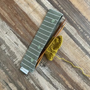 Peut inclure: Un étui à aiguilles en tissu rayé gris et blanc avec trois aiguilles à tricoter en bois et un petit morceau de tricot jaune. L'étui est doté de trois boutons-pression et le texte "chinashu" est imprimé en bas.