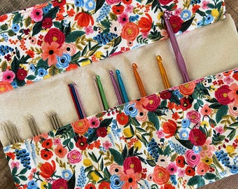 Puede incluir: Un organizador de tela enrollable con un vibrante estampado floral de flores rojas, rosas, naranjas y azules sobre fondo blanco, que contiene varios ganchillos de metal en colores variados, incluyendo morado, verde y dorado.