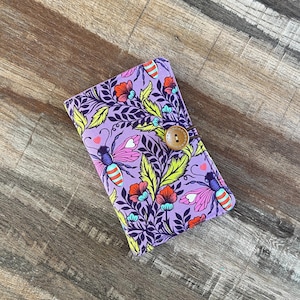 May include: A purple fabric wallet featuring a floral and bee design. The pattern includes red, pink, and blue flowers, green leaves, and stylized bees. A wooden button fastens the wallet.