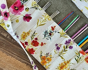 Puede incluir: Un estuche de tela para ganchillos con un estampado floral de flores rojas, rosas, amarillas y moradas sobre fondo blanco. El estuche cuenta con un cierre de botón a presión morado y contiene varios ganchillos de metal y aluminio de colores.