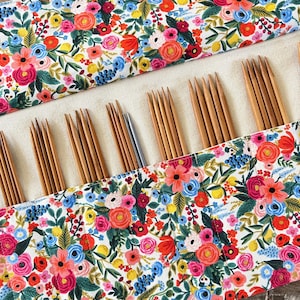 Könnte beinhalten: Eine Stricknadel-Tasche mit floralem Muster und Stricknadeln aus Holz. Die Tasche hat einen weißen Hintergrund mit einem farbenfrohen Muster aus rosa, roten, blauen und gelben Blumen und grünen Blättern. Die Nadeln sind in verschiedenen Größen und aus Holz.