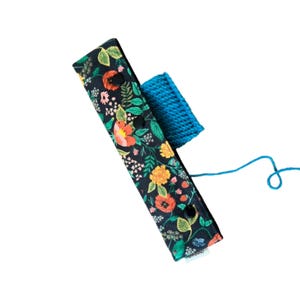 May include: A floral print yarn swift with a blue knitted yarn holder. The fabric is black with a colorful floral pattern of orange, yellow, and green flowers. The yarn is blue.