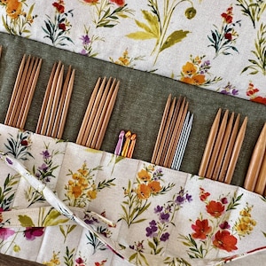 May include: A floral print knitting needle case with various wooden and metal knitting needles. The case is made of a cream-colored fabric with a colorful floral pattern. The needles are of different sizes and materials, perfect for knitting projects.