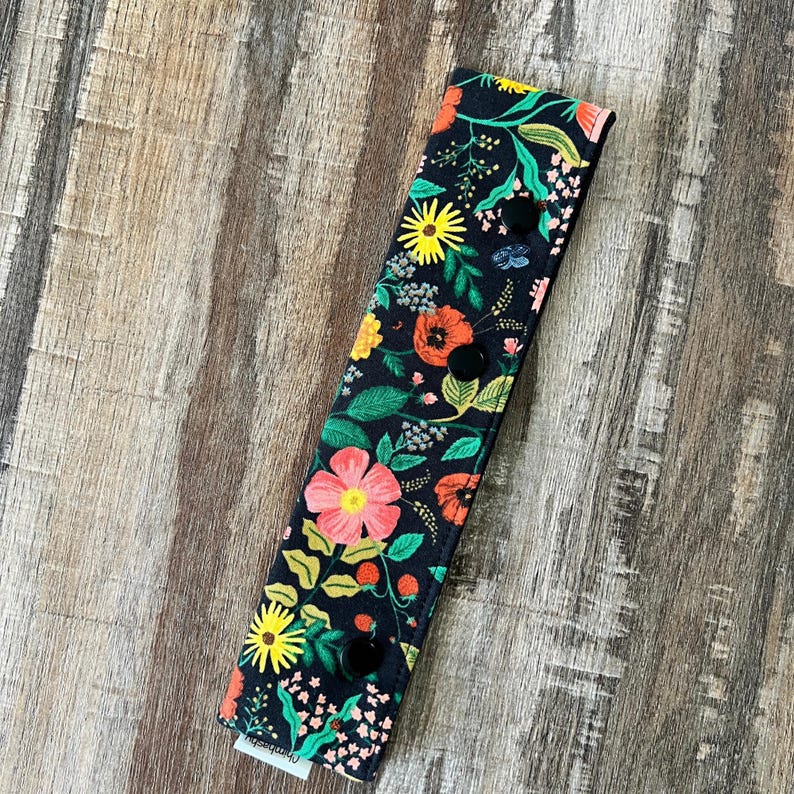 May include: A black floral print headband with yellow, orange, and pink flowers, green leaves, and three black buttons. The headband is rectangular and made of fabric.