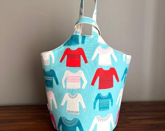 Puede incluir: Un bolso tote de color azul claro con un estampado repetido de suéteres rojos, blancos, rosas y turquesas. El bolso tiene un asa a juego y un diseño abierto, perfecto para llevar objetos.